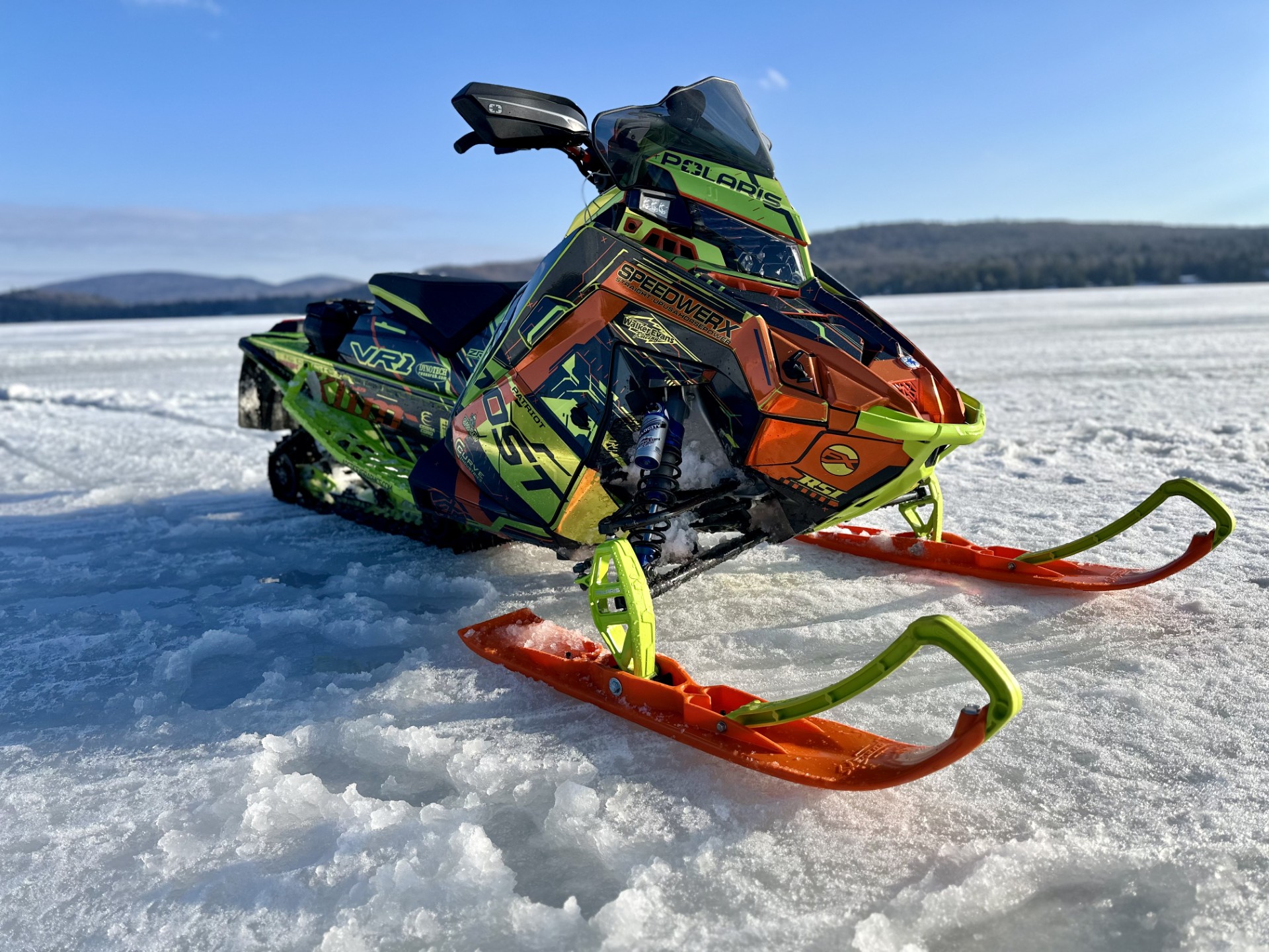 A Polaris Matryx Trail snowmobile with a gunmetal gray, orange, and hyper green KLIM Recoil wrap with chrome vinyl and halo flake laminate.