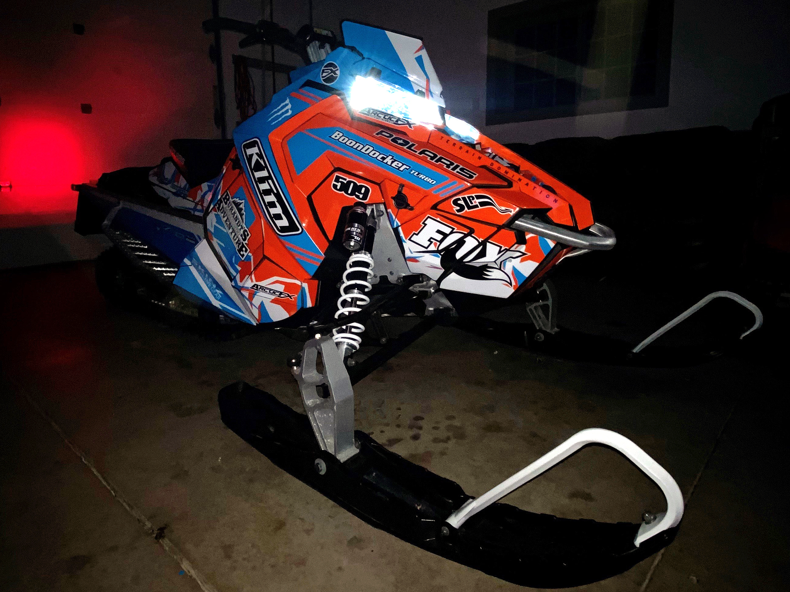 A Polaris Axys Mountain snowmobile with a blue, orange, and white Burandt Vista custom vinyl wrap.