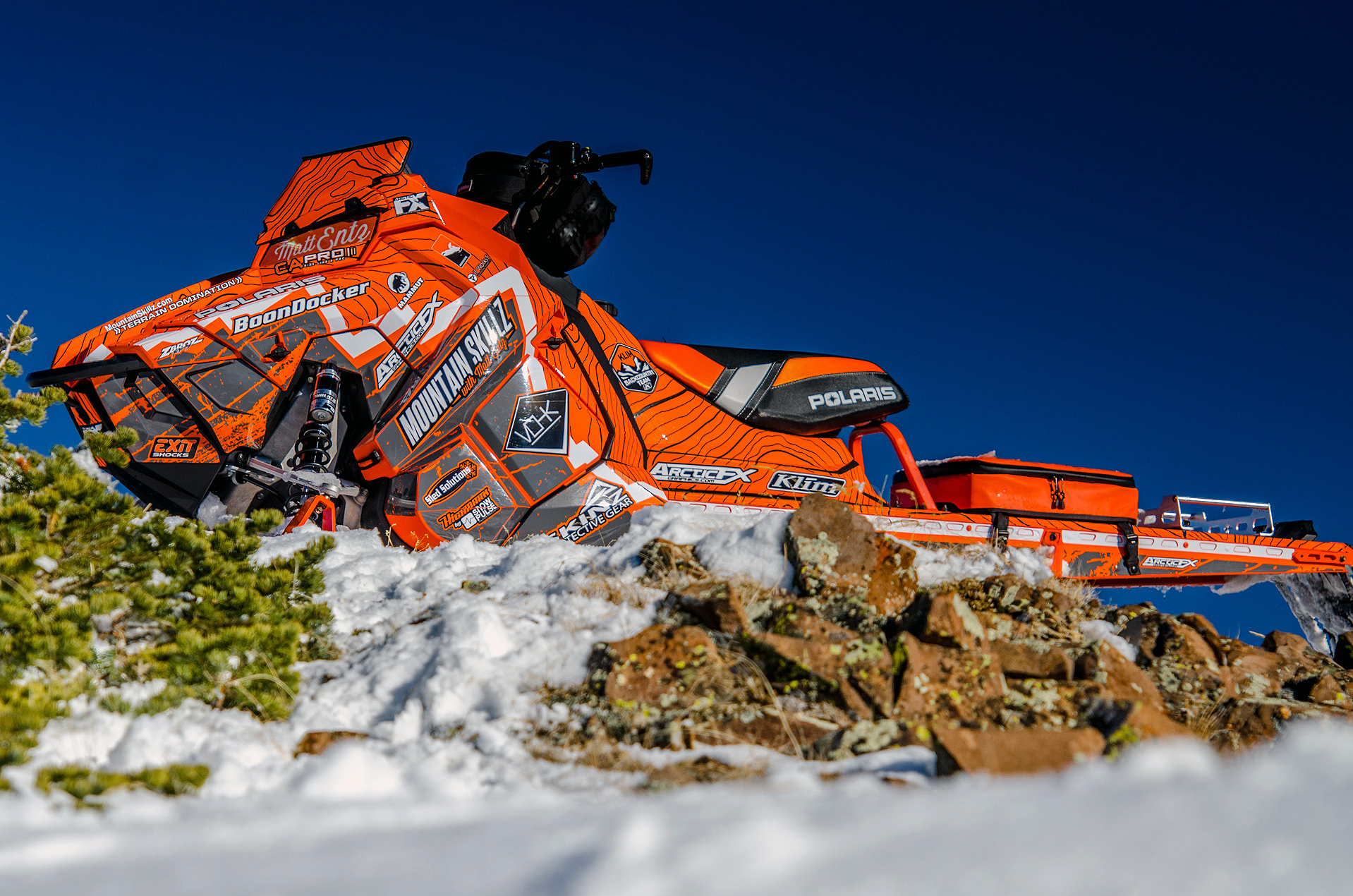 A Polaris Axys Mountain snowmobile with a orange, gray, and white Entz Frontier custom vinyl wrap.