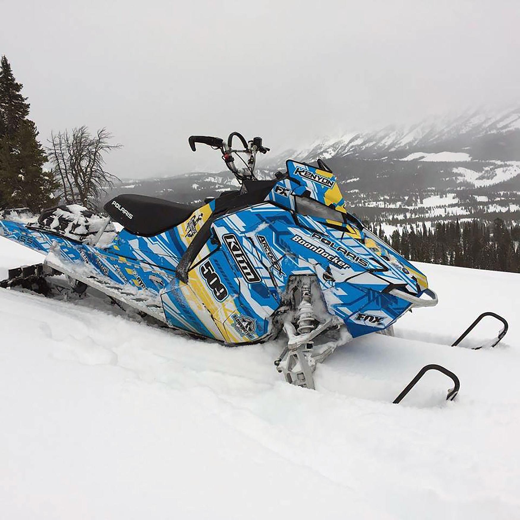 A Polaris Axys Mountain snowmobile with a blue, yellow, and white Evasion custom vinyl wrap.