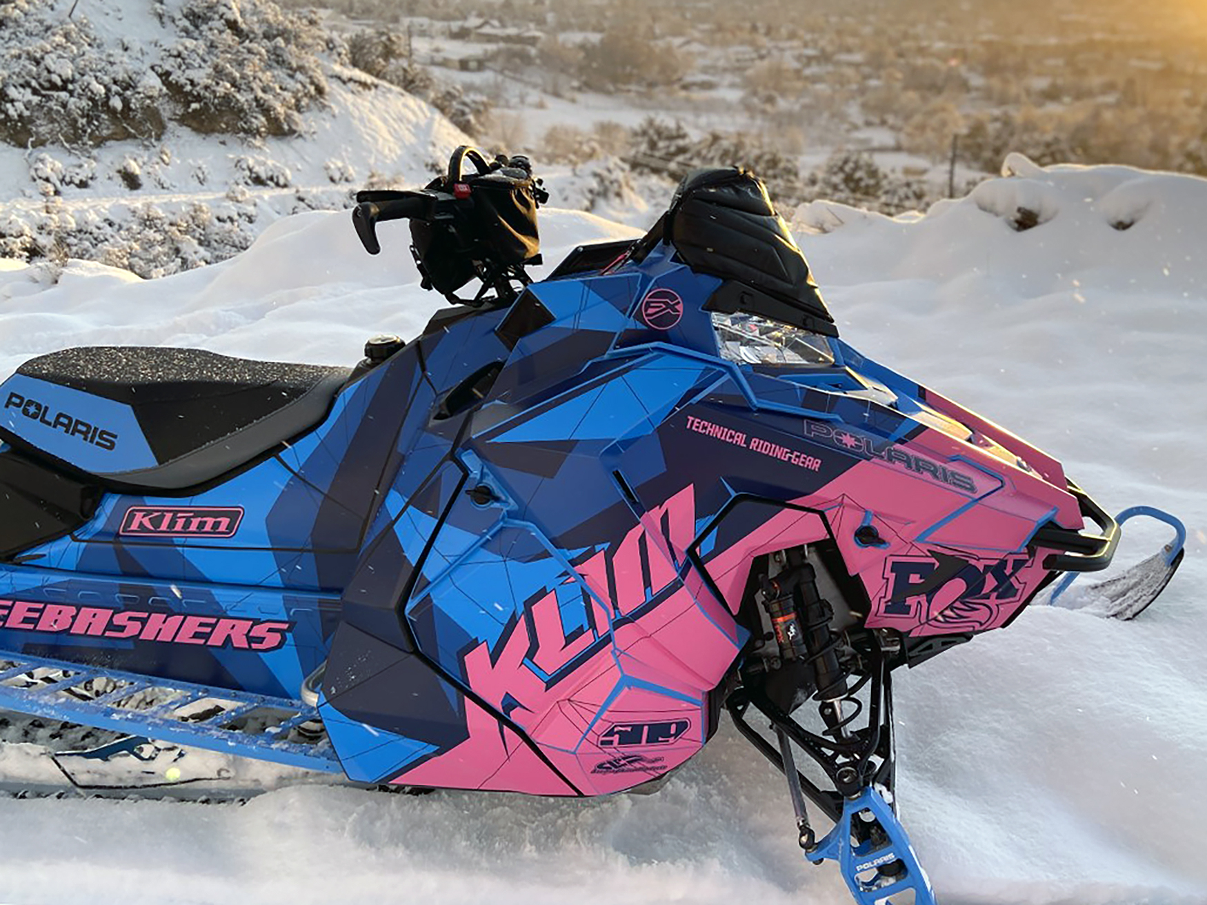 Two Polaris Axys Mountain snowmobiles with a pink, blue, and black Klim F3 Camo custom vinyl wraps.