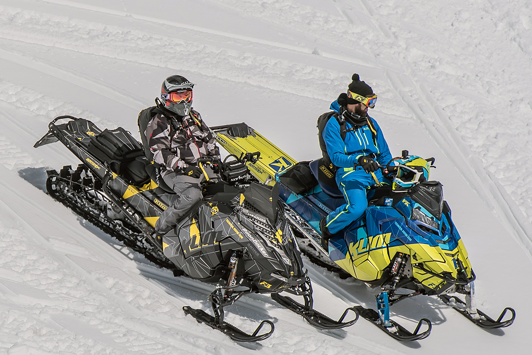 Two Polaris Axys Mountain snowmobiles with a blue and gray and lime squeeze Klim F3 Camo custom vinyl wraps.