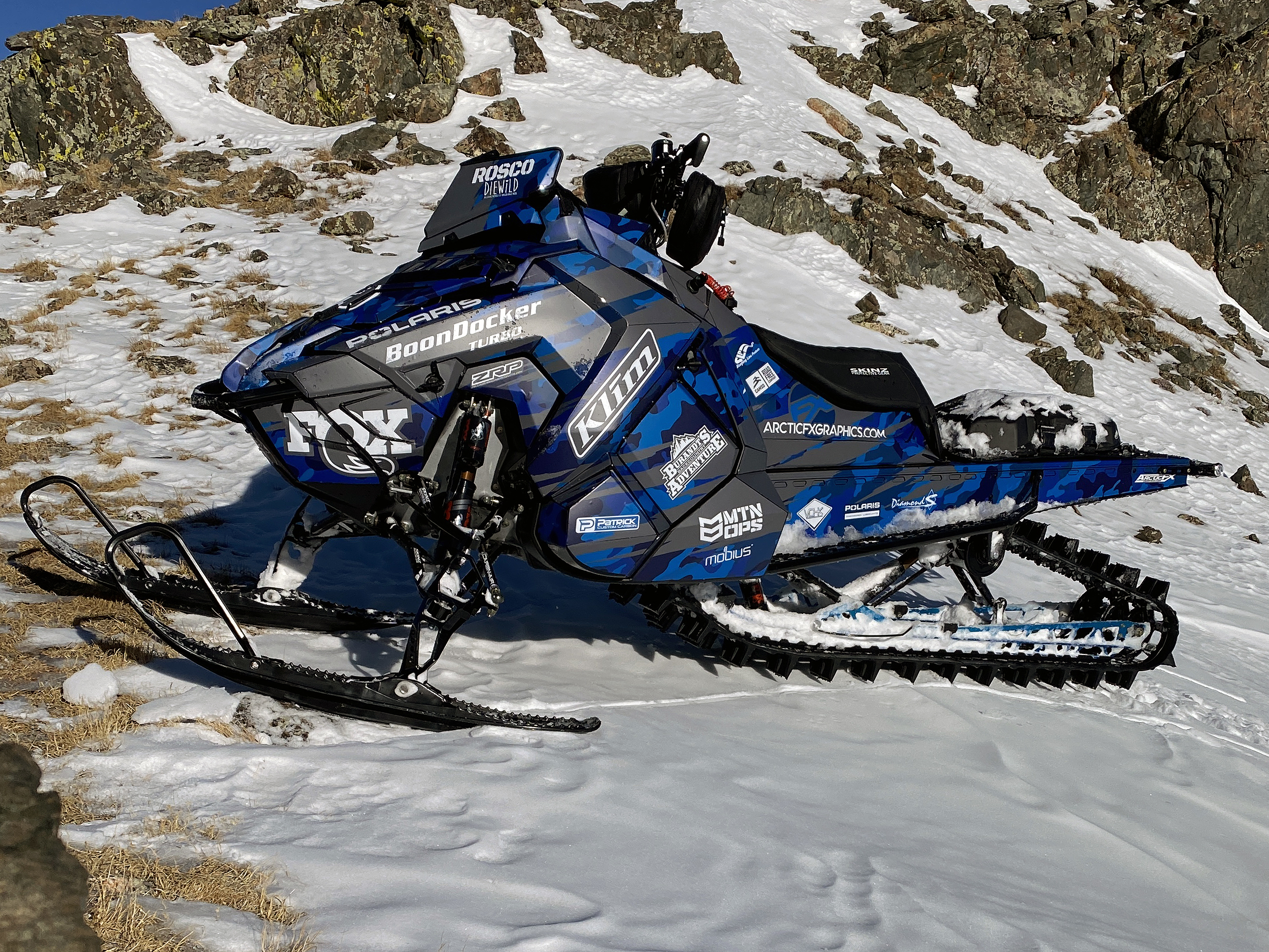 A Polaris Axys Mountain snowmobile with a blue, black, and gray RR Camo custom vinyl wrap.
