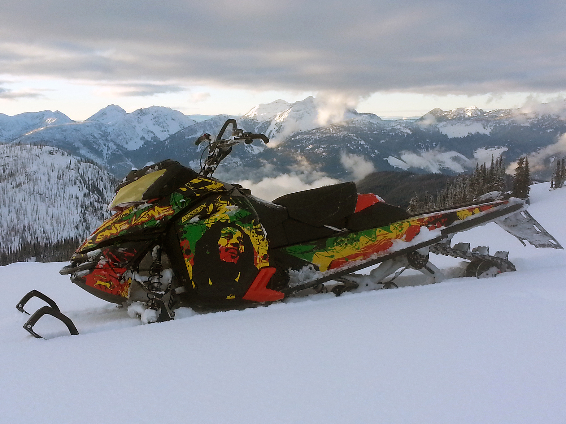 A Ski-Doo Rev XM snowmobile with a yellow, green, red, and black rasta Bob Marley custom vinyl wrap.