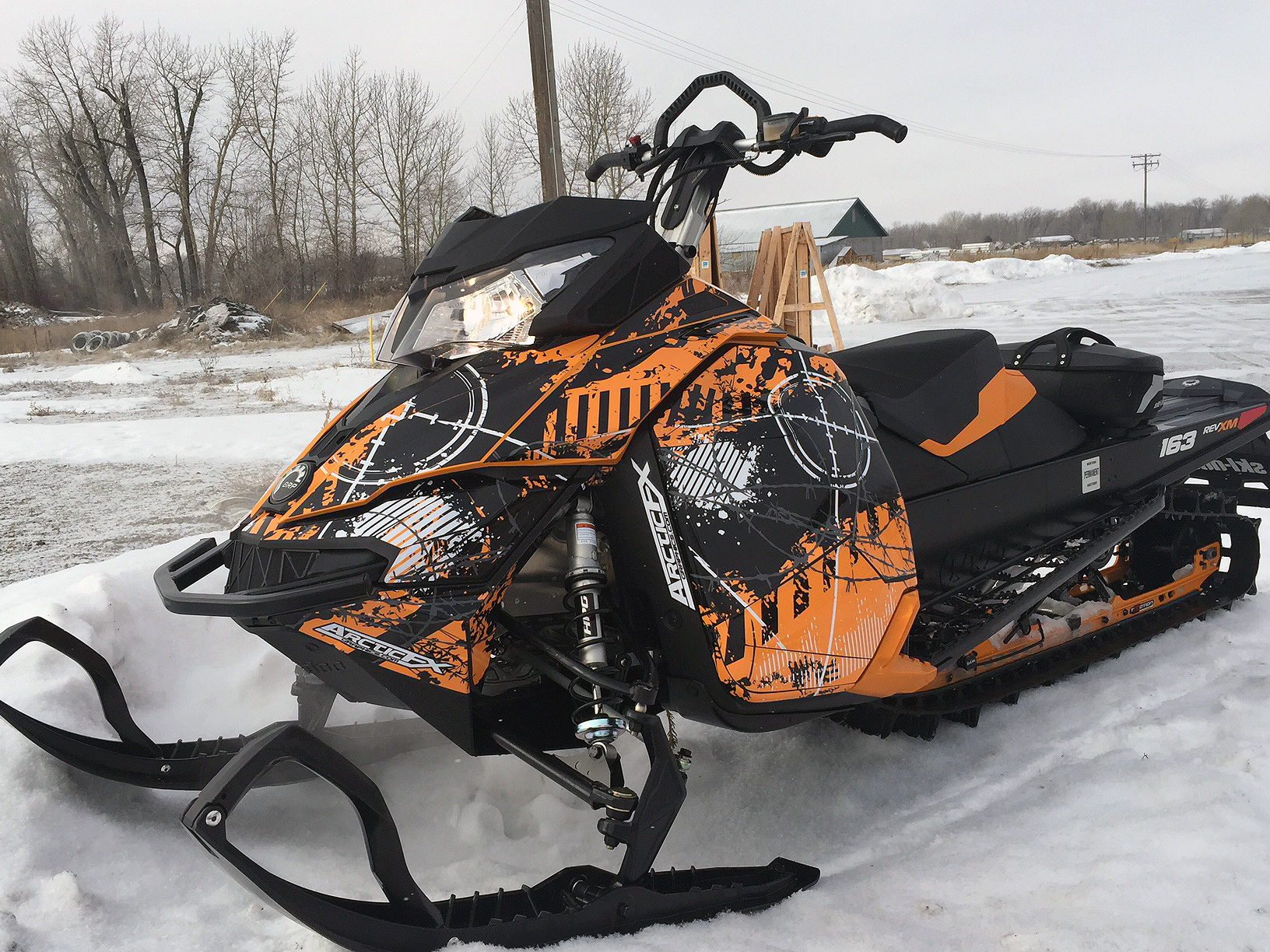A Ski-Doo Rev XM snowmobile with a orange, white, and black Bombsquad custom vinyl wrap.