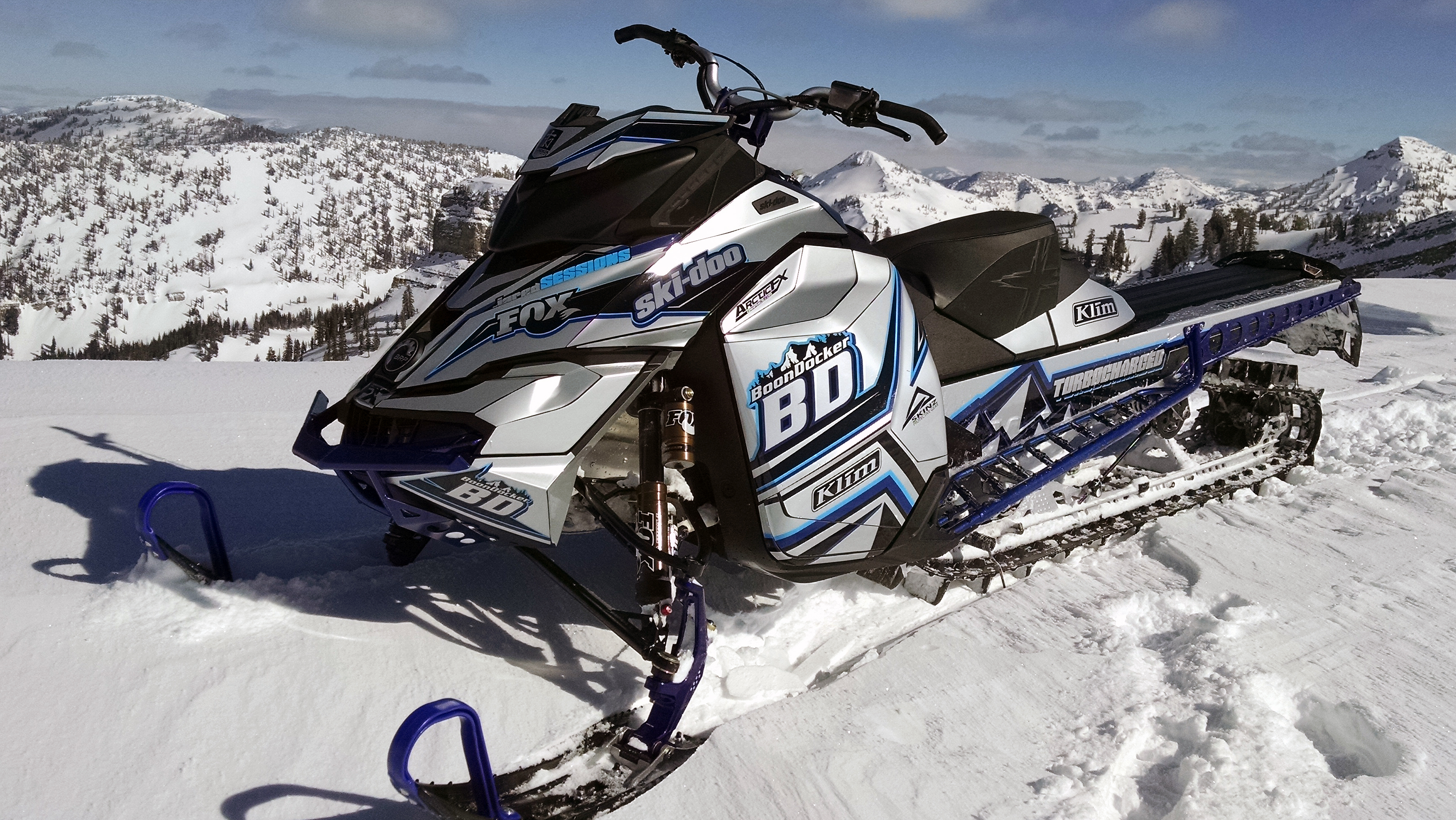 A Ski-Doo Rev XM snowmobile with a gray, blue, and white Sessions custom vinyl wrap.