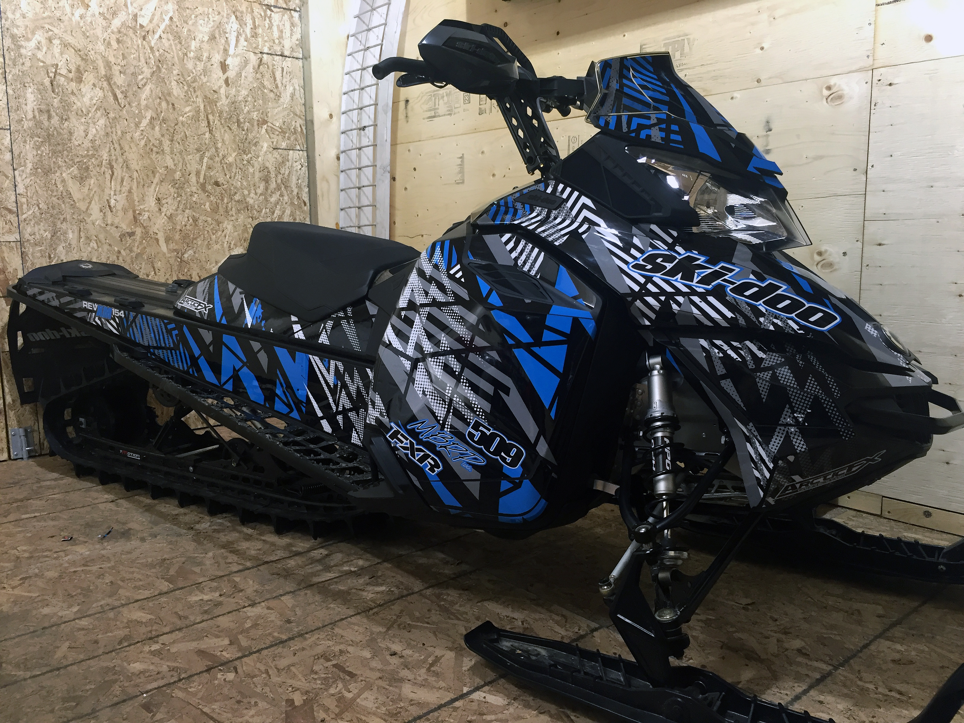 A Ski-Doo Rev XM snowmobile with a blue, white, and black Voltage custom vinyl wrap.