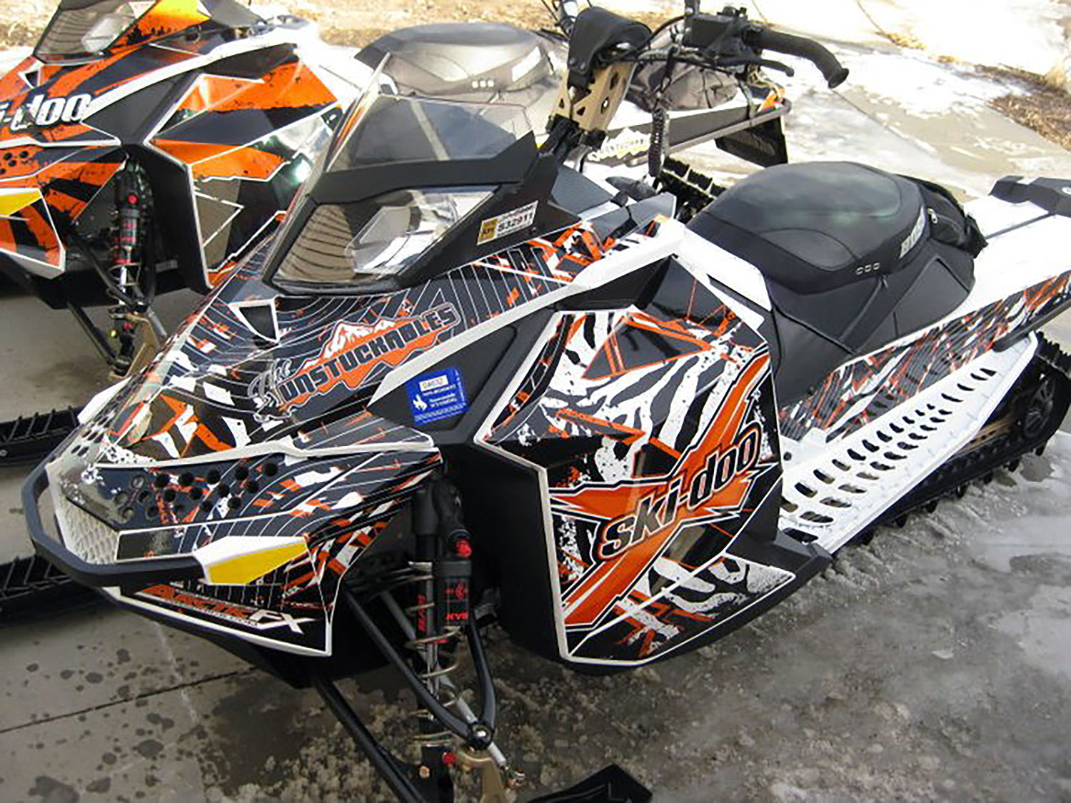 A Ski-Doo Rev XP snowmobile with a black, white, and orange Carnivore custom vinyl wrap.