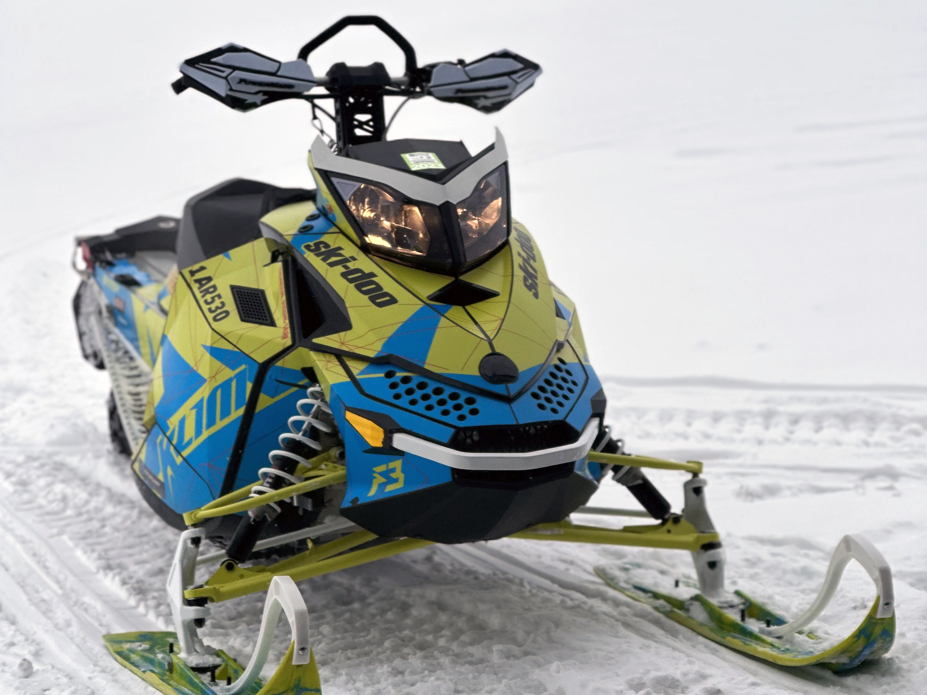 A Ski-Doo Rev XP snowmobile with a yellow, blue, and red Klim F3 Camo custom vinyl wrap.
