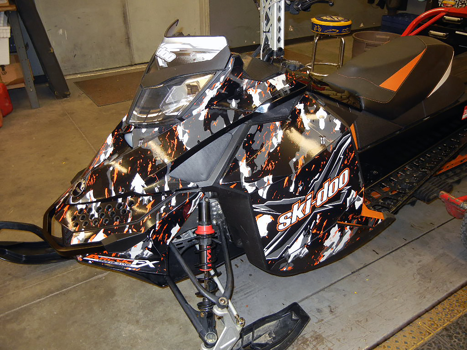 A Ski-Doo Rev XP snowmobile with a black, gray, and orange Urban Camo custom vinyl wrap.