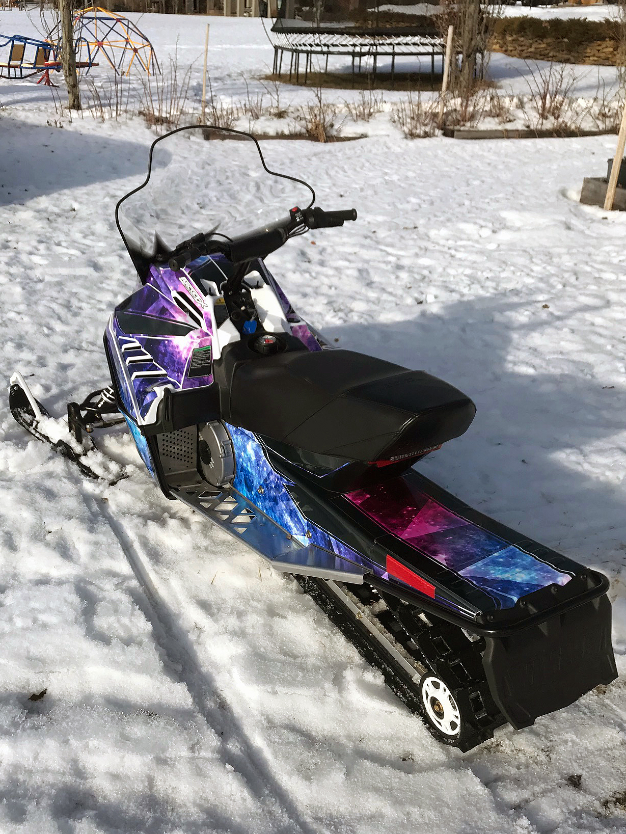 A Yamaha SnoScoot youth snowmobile with a black, purple, and blue galaxy Cosmic Camo custom vinyl wrap.